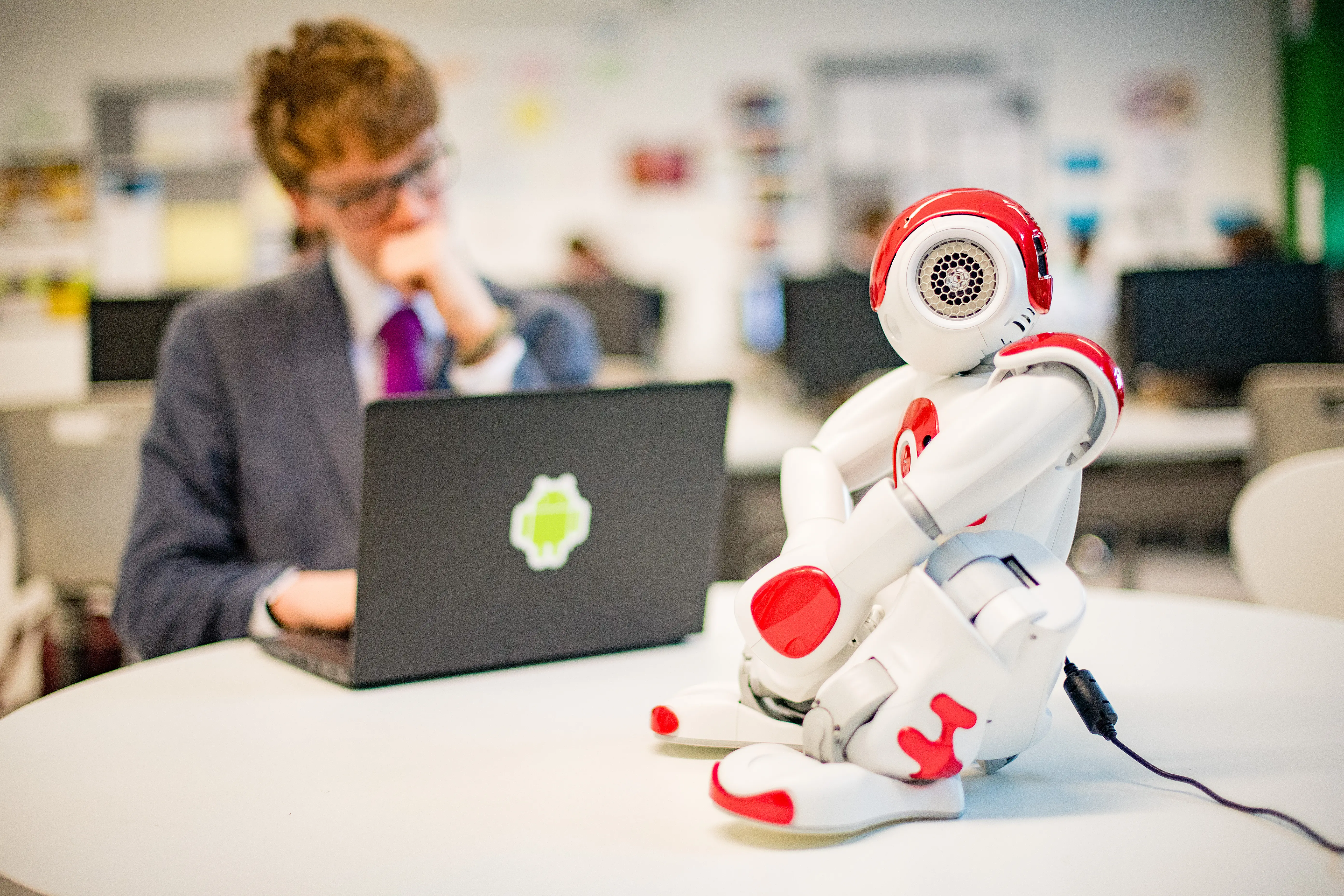 A small humanoid robot on a desk beside someone working on a laptop